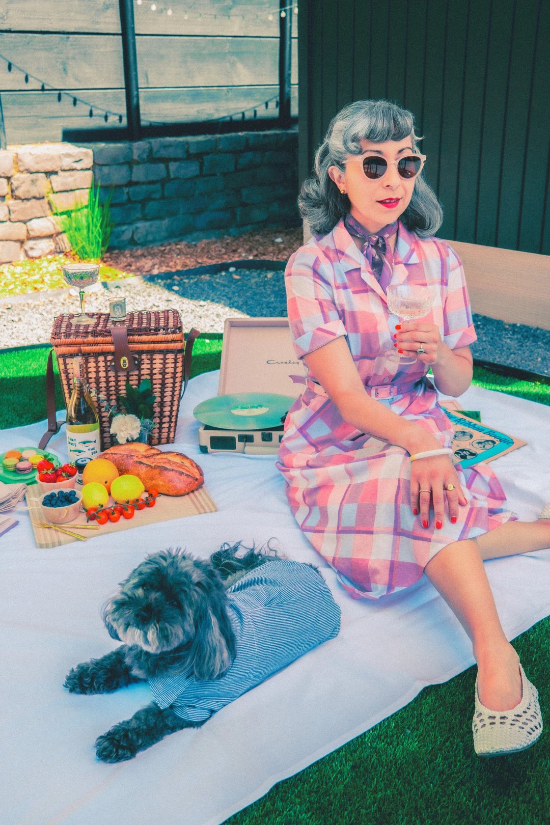 A woman in a pink checkered dress holds a glass of drink while sitting on a picnic blanket surrounded by food, a picnic basket, wine, and a record player.