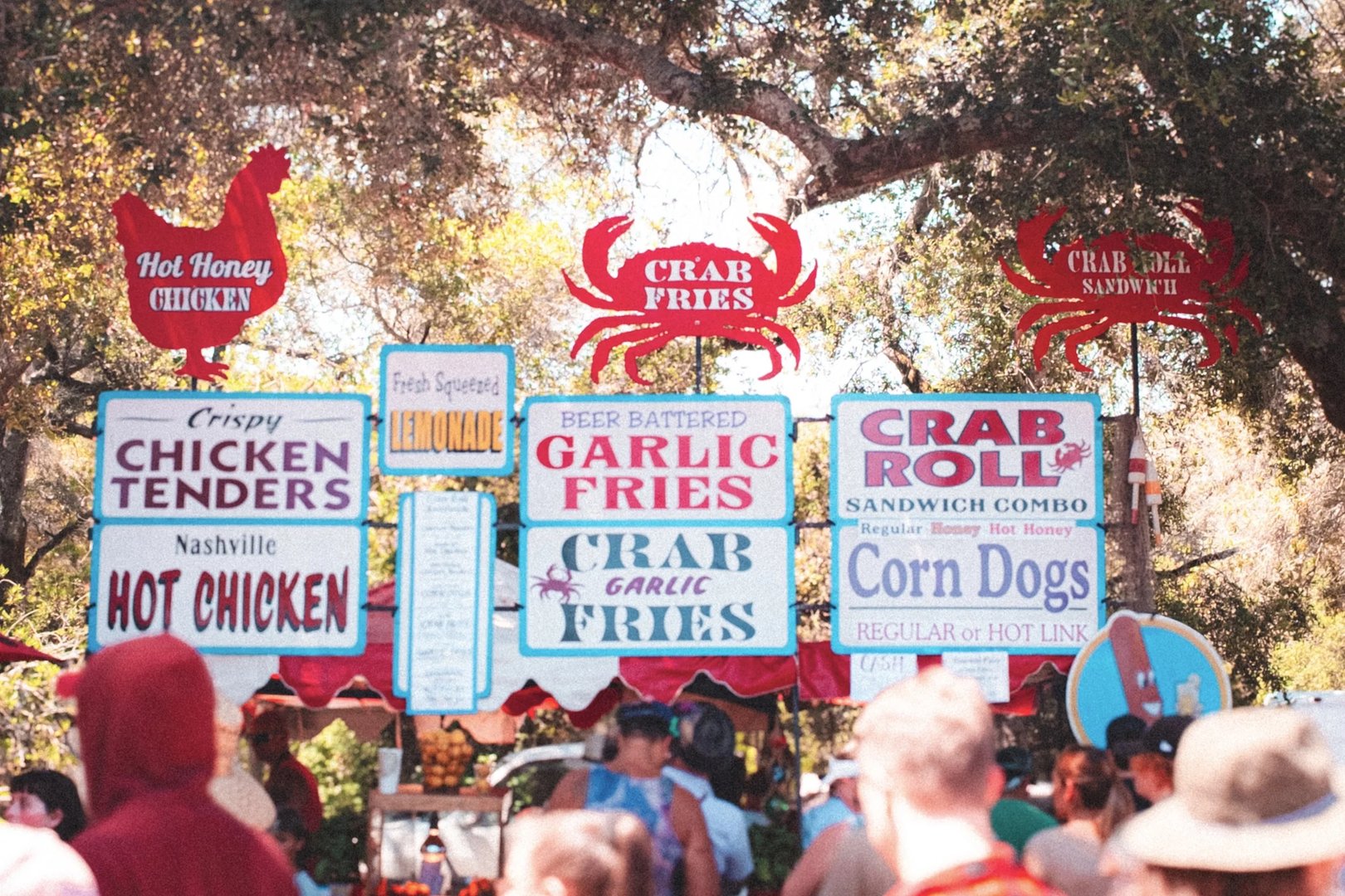A food stand with signs advertising various items including crab fries, chicken tenders, and corn dogs in a crowded outdoor setting.