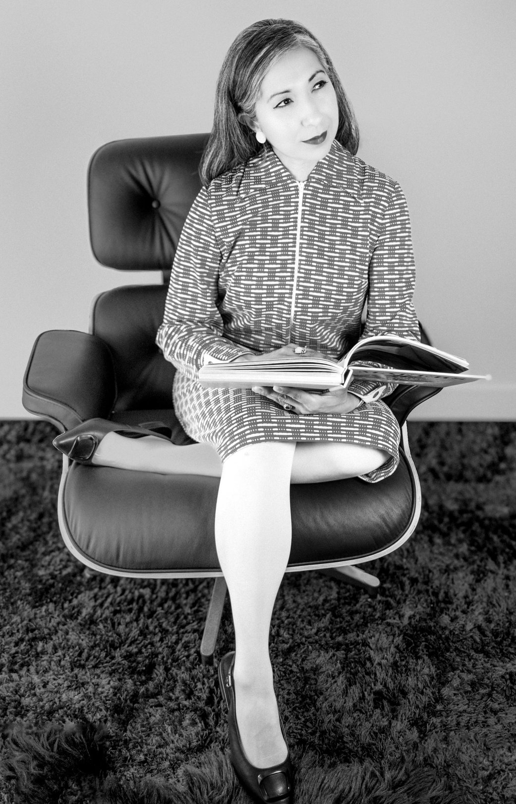 A person sitting in a vintage chair, reading a magazine, dressed in a patterned outfit with a minimalist background.