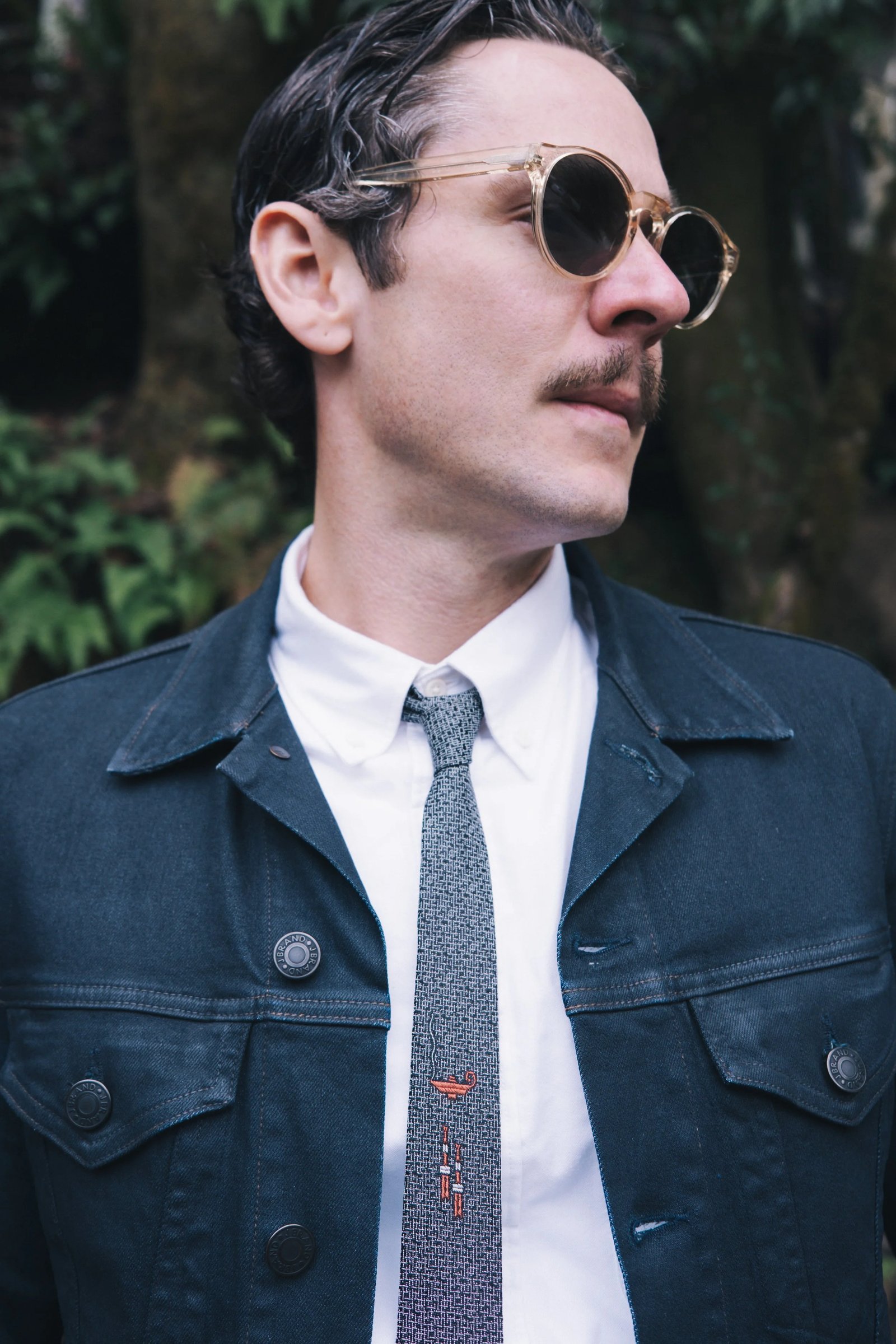 A man wearing sunglasses and a tie stands confidently in a dark jacket against a green backdrop.