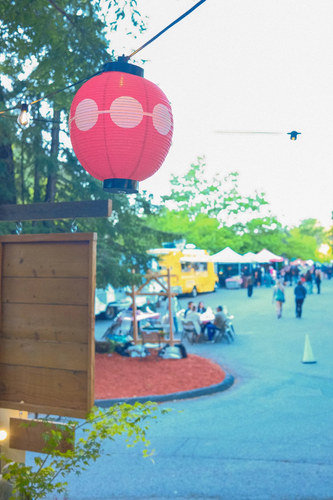 A red lantern with white dots hangs above a busy outdoor marketplace featuring food trucks and people sitting at tables.