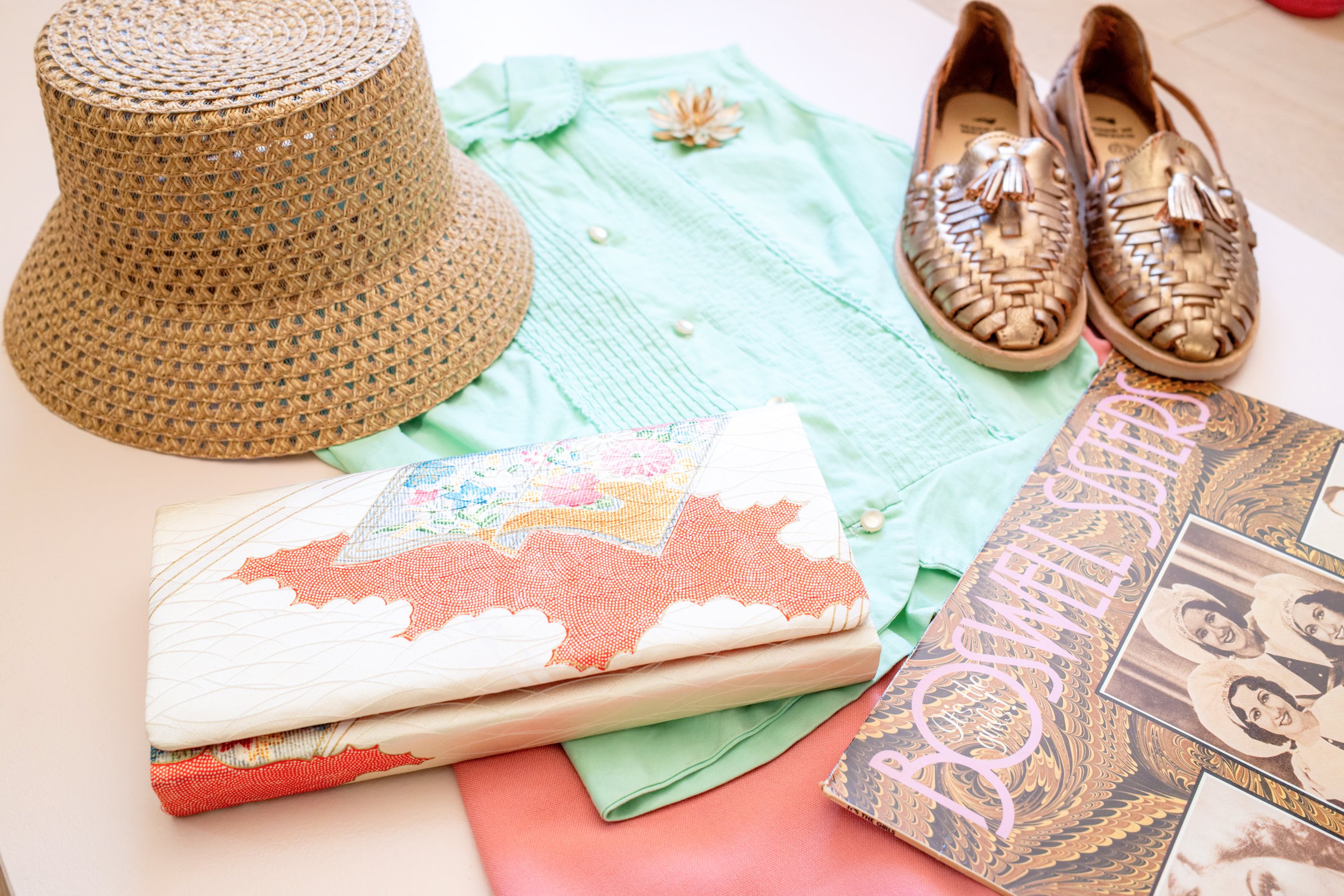 Inspired by Summer, Styled by Music gallery image: A woven straw hat, a green button-up shirt with decorative stitching, a pair of metallic brown shoes, a floral-patterned clutch, and a vintage magazine titled "Boswell Sisters.