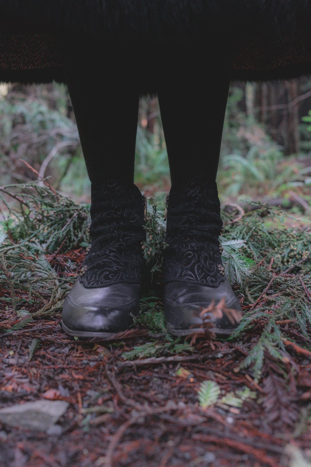 A person stands on forest floor wearing black shoes and decorative black socks amidst greenery.