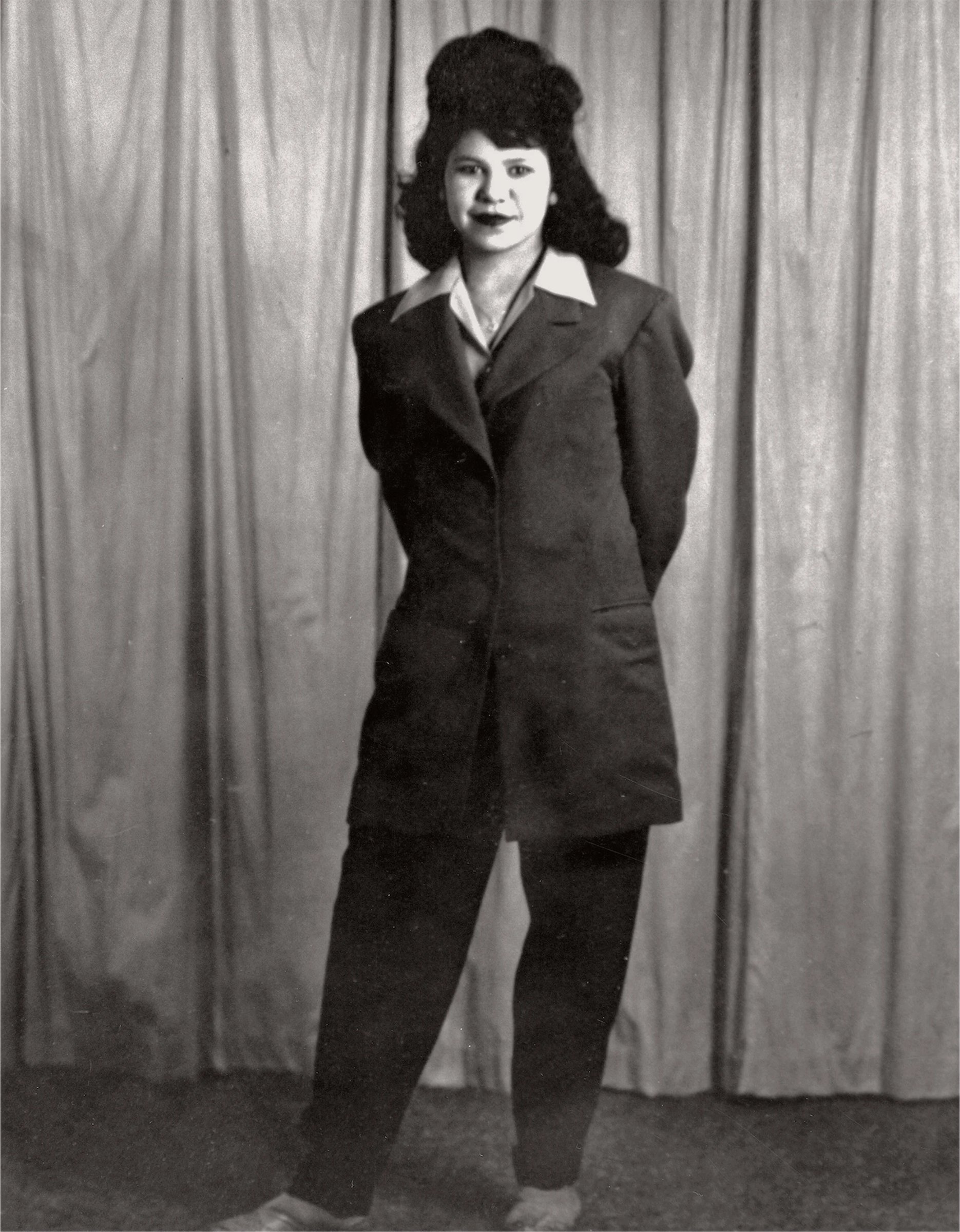 A woman poses standing in a vintage suit with a high collar and a large hairstyle against a curtain backdrop.