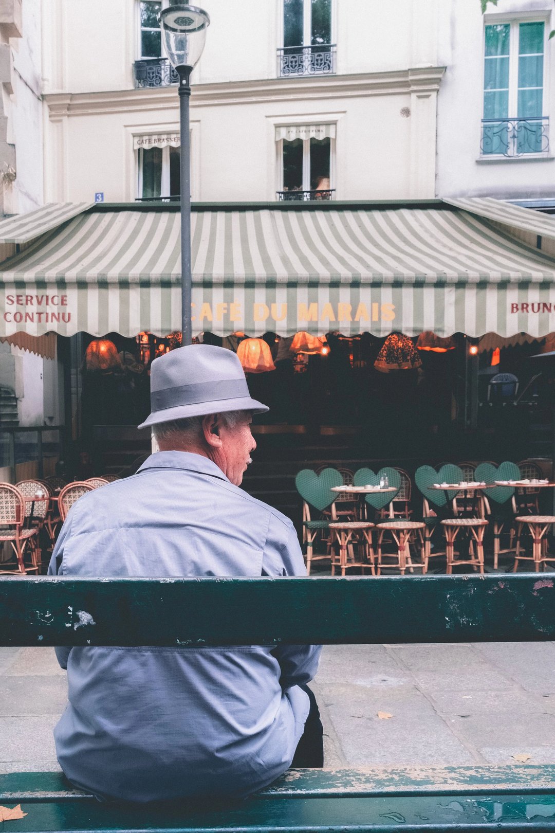 A man wearing a gray hat sits on a bench in front of a cafe with striped awnings and empty chairs.
