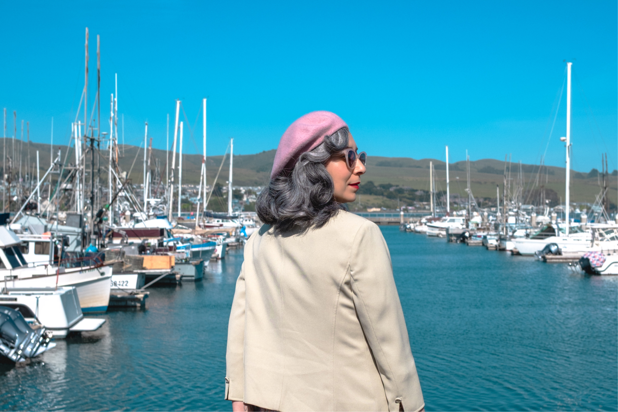The Road to Bodega Bay gallery image: A woman in a pink beret and sunglasses stands by a marina filled with boats, facing away from the camera against a clear blue sky.