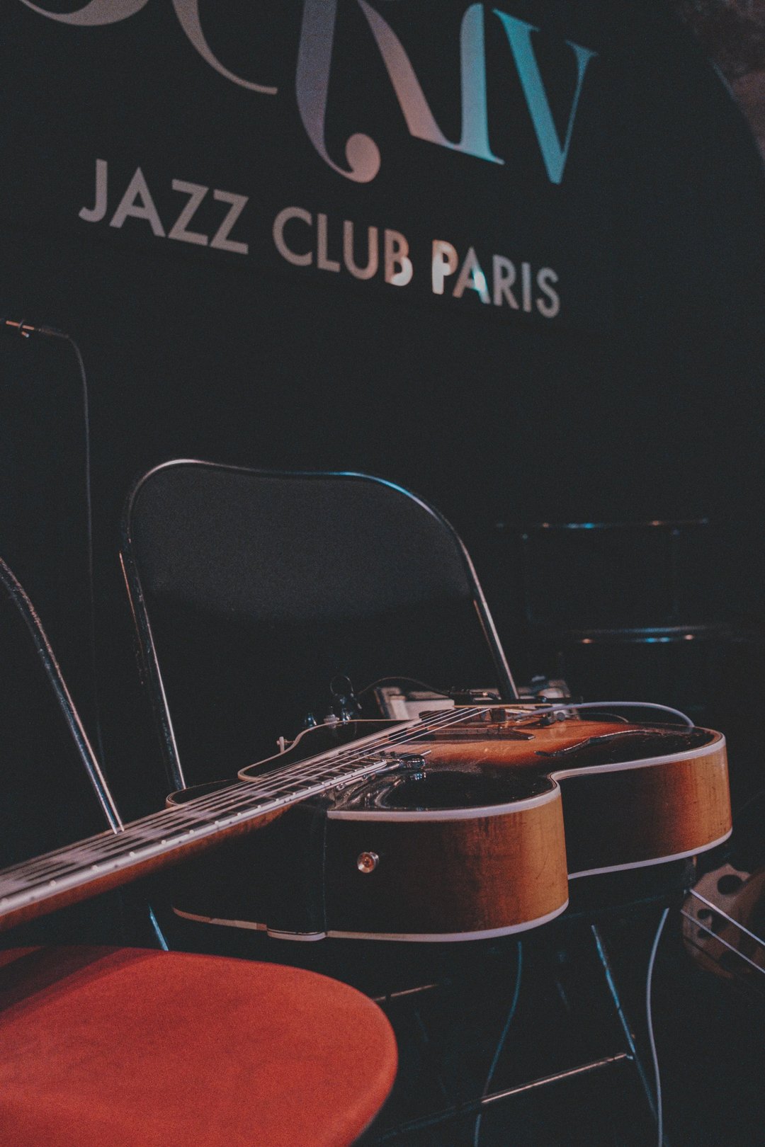 A close-up of a guitar resting on a chair in a jazz club with a sign that reads "Jazz Club Paris" in the background.