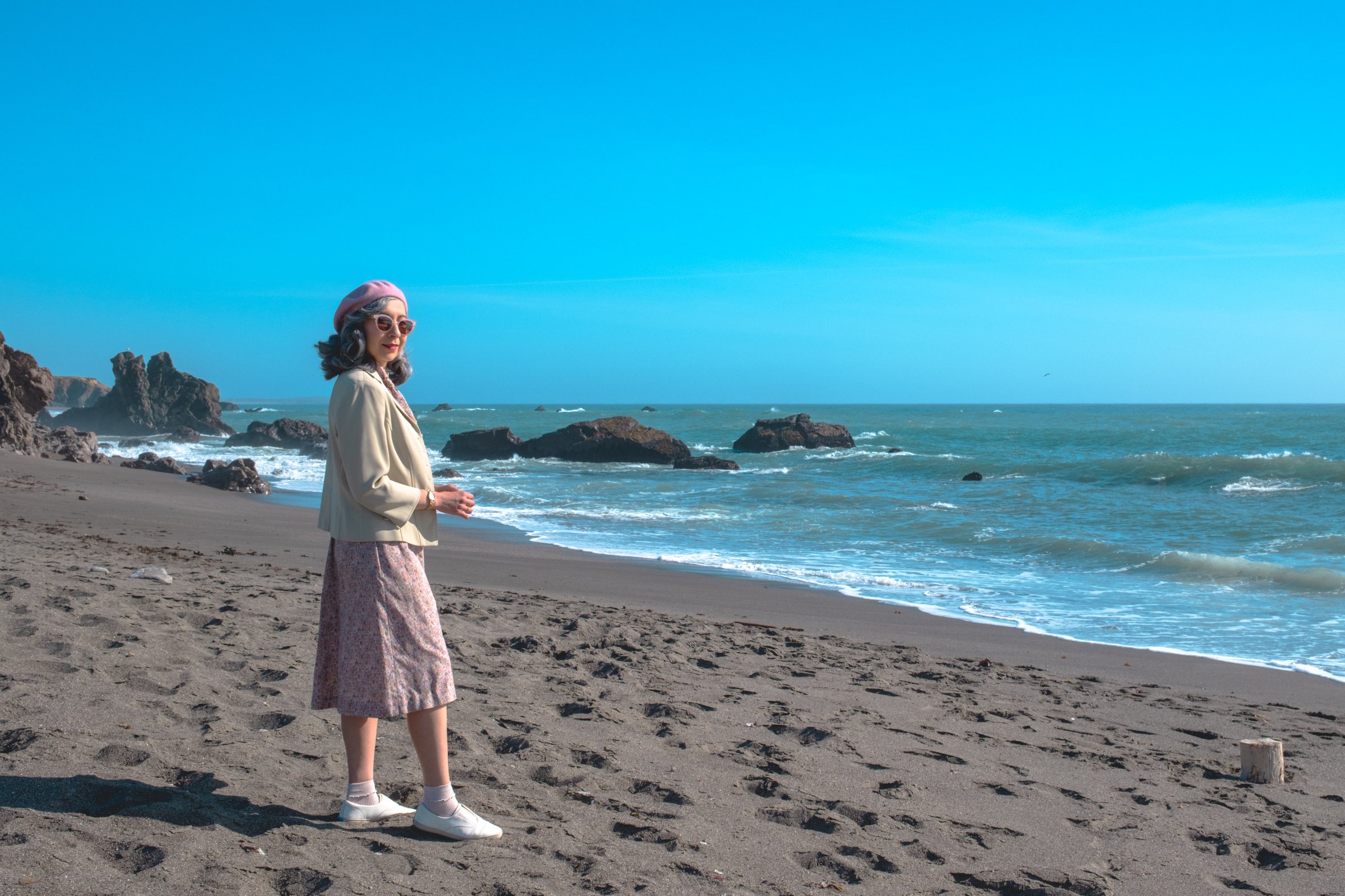 The Road to Bodega Bay gallery image: A woman in a light jacket, pink beret, and patterned skirt stands on a sandy beach with waves and rocks in the background under a clear blue sky.