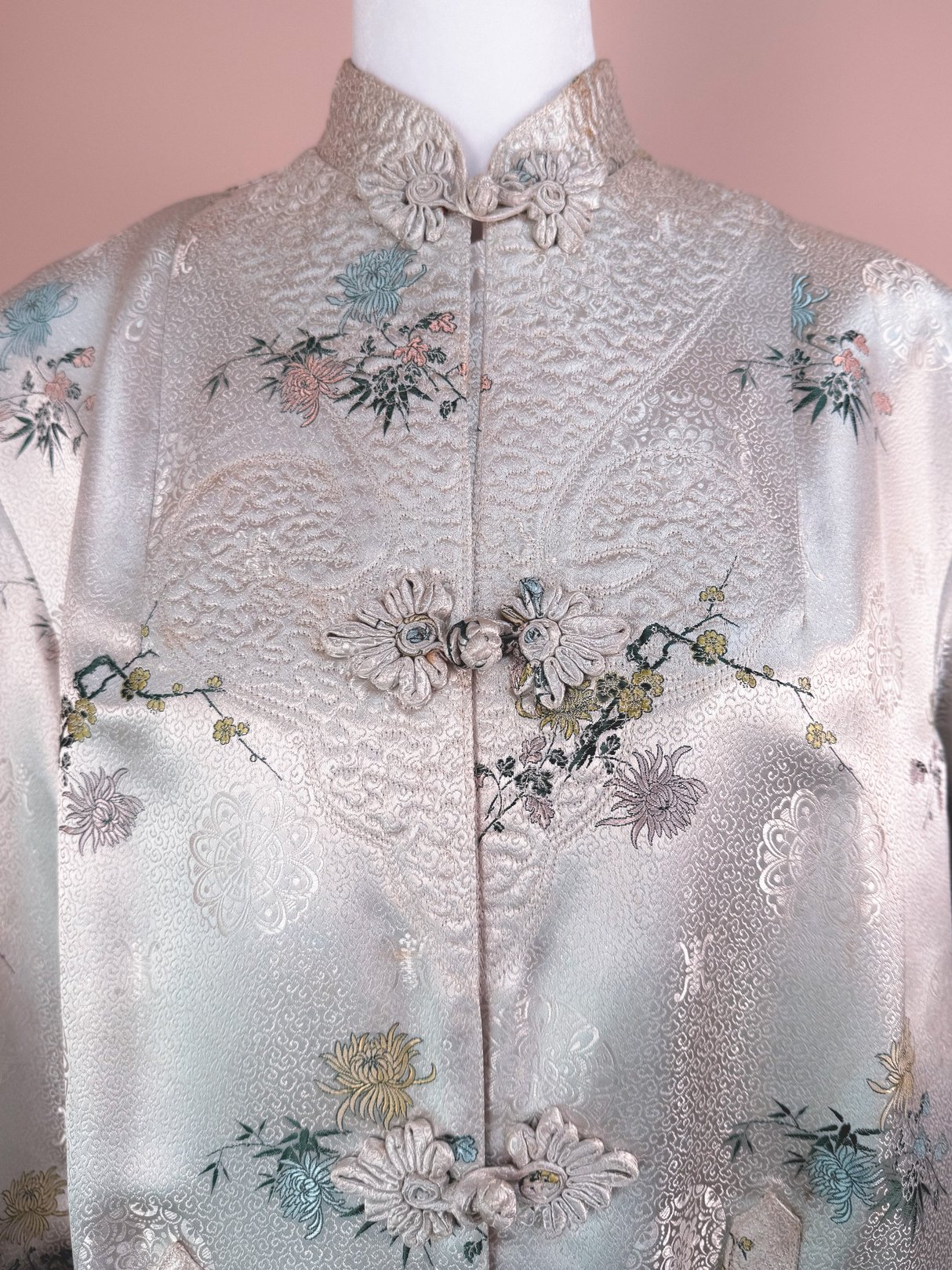 Silver silk cheongsam coat for women featuring intricate floral embroidery and decorative buttons, representative of 1970s design.