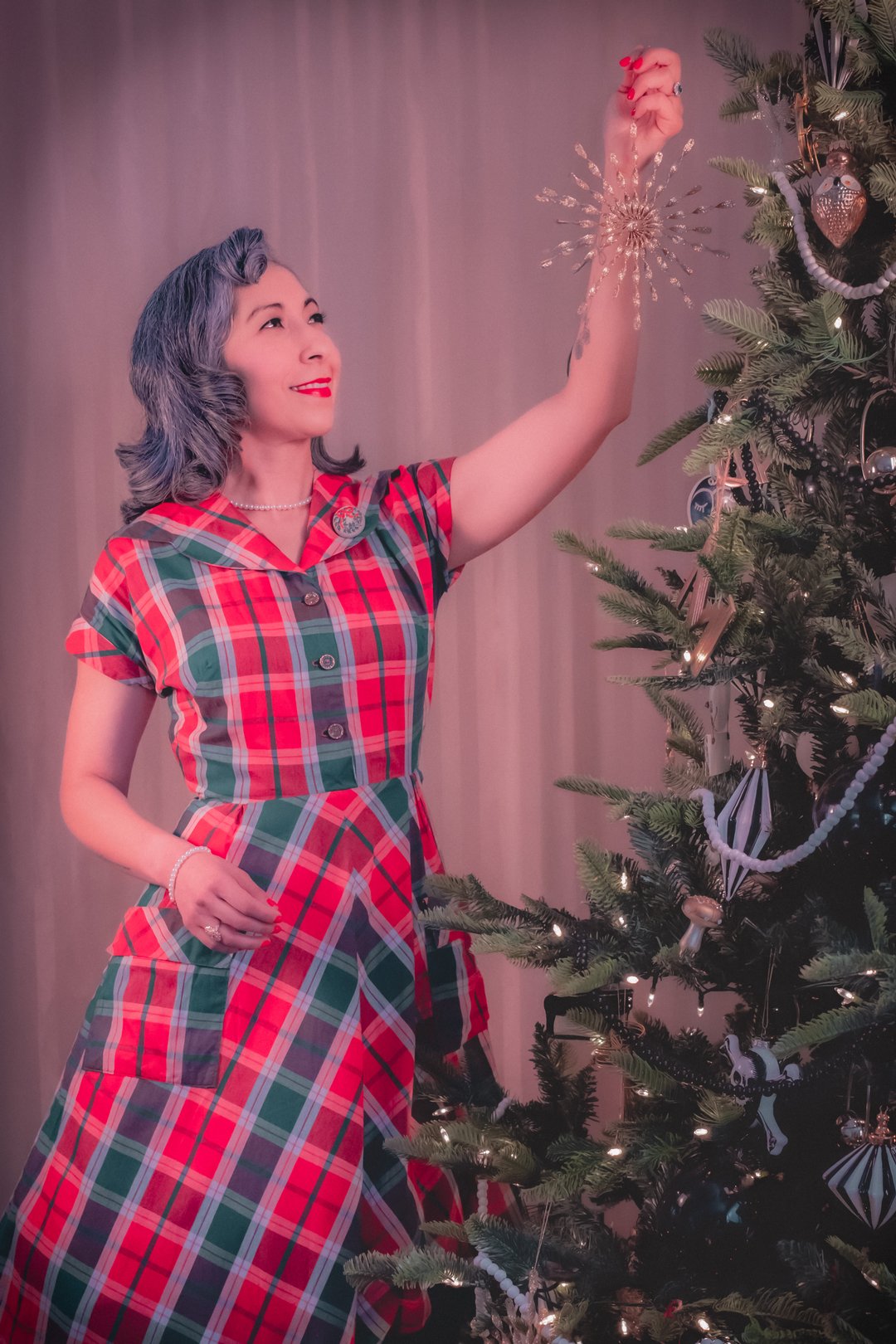 A woman in a red and green plaid dress decorates a Christmas tree with a snowflake ornament.