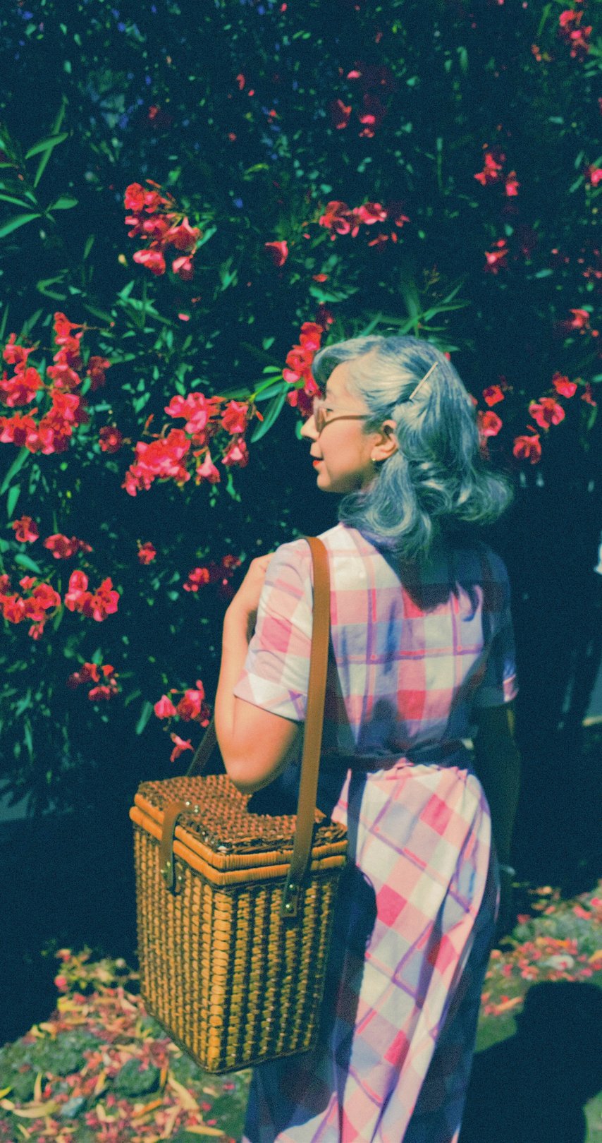 A woman with pastel blue hair stands amidst vibrant pink flowers, wearing a checkered shirt and carrying a brown shoulder bag.