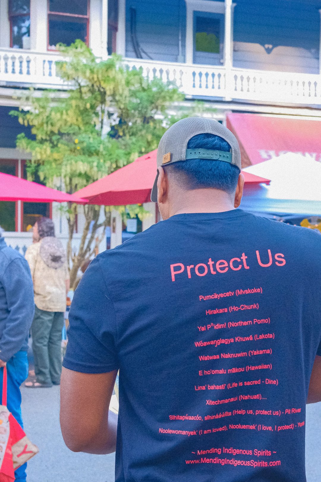 A person wearing a black t-shirt with the phrase "Protect Us" and various Indigenous names and languages listed on the back.