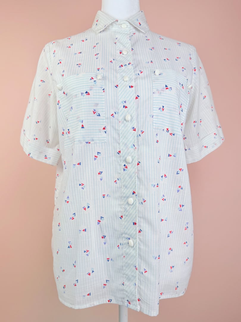 White short-sleeve top with blue stripes and red and blue flag patterns, featuring a button-down front and two chest pockets, typical of 1980s women's fashion.