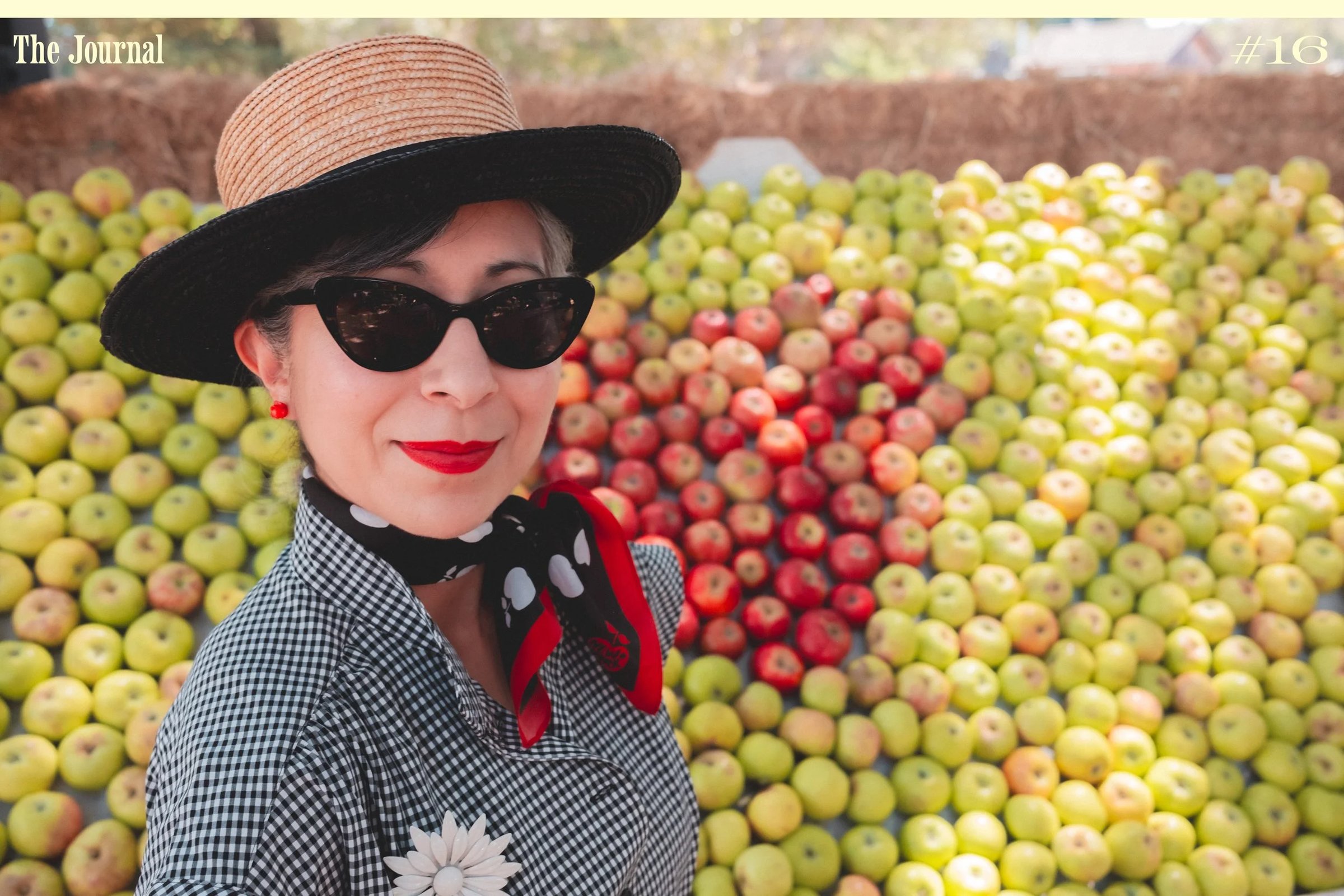 A woman wearing a straw hat and sunglasses stands in front of a colorful display of apples at a market.
