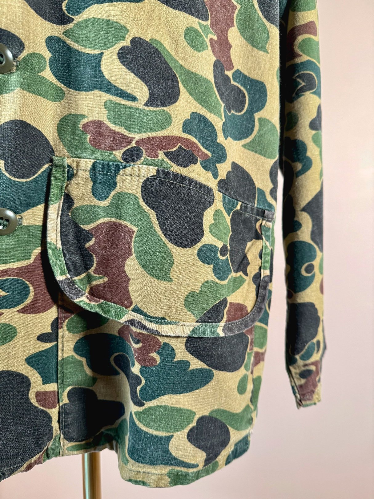 Camo jacket in camouflage green featuring a button closure and front pockets, styled for men, reflecting 1970s fashion.