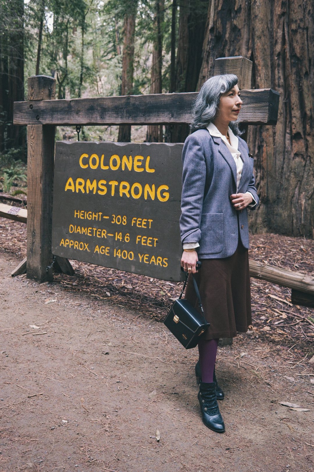 A woman stands beside a sign for Colonel Armstrong, a 308-foot tall, 1,400-year-old tree in a redwood forest.