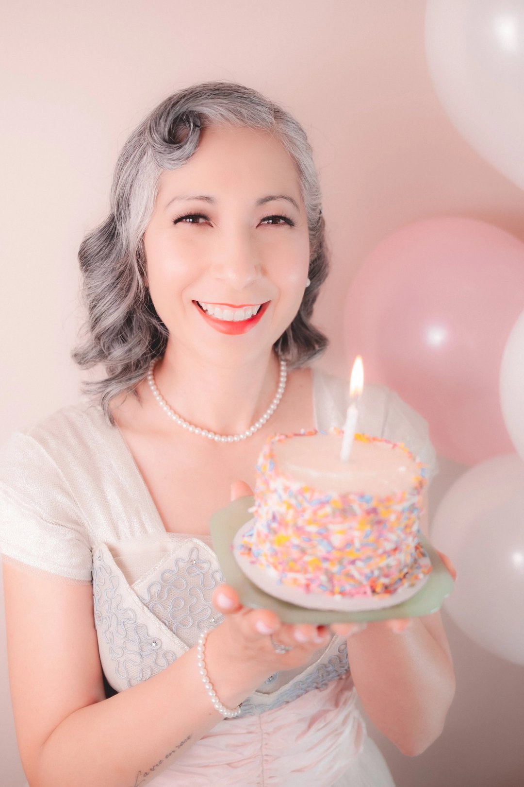 A woman with gray hair holds a decorated birthday cake with a single candle in front of pastel balloons.