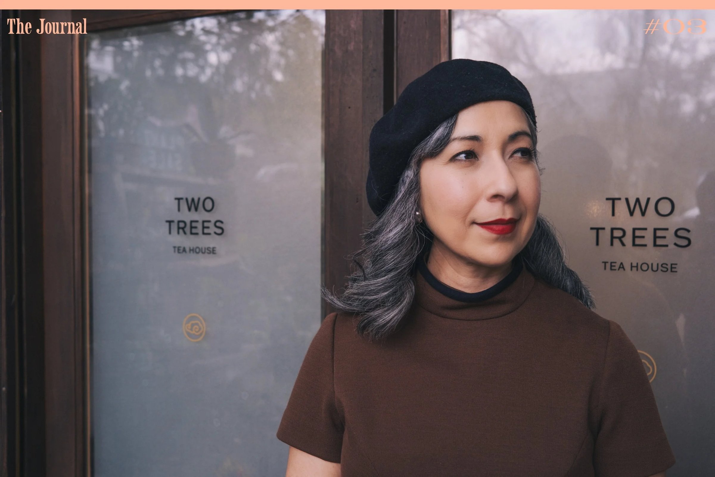 A woman with gray hair and a black beret stands in front of a wooden door with the text "Two Trees Tea House.