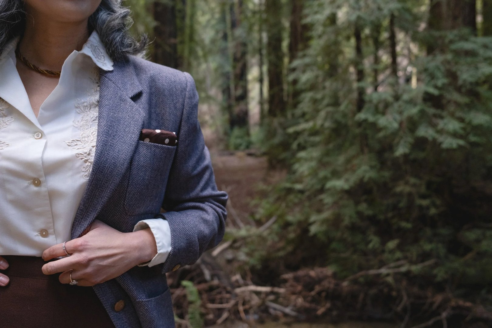 A person wearing a herringbone blazer stands in a forest setting, holding a small item in their hand.