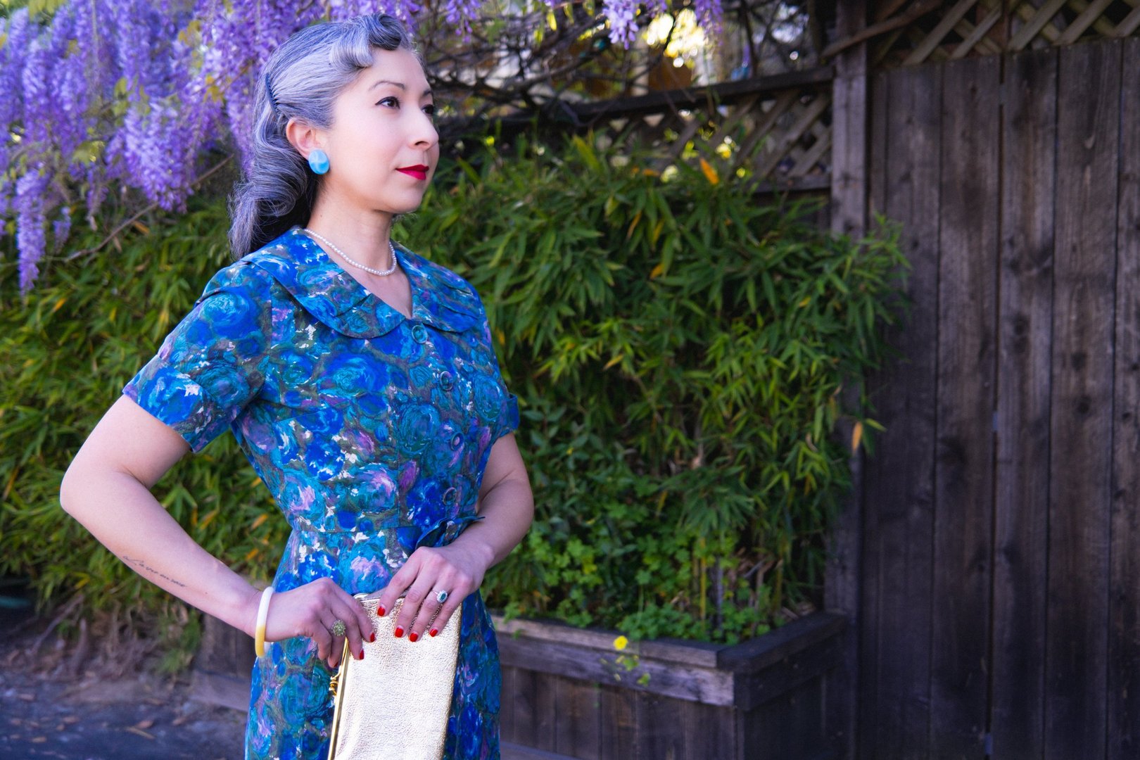 A woman wearing a 1950s blue floral skirt set stands outdoors, holding a gold clutch, with wisteria in the background.