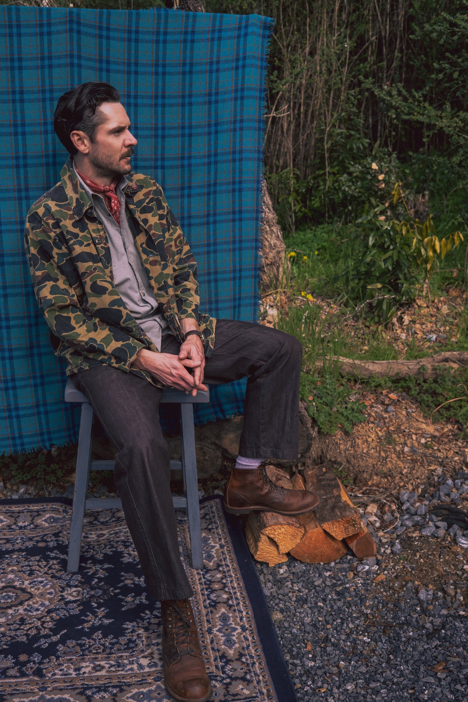 A person sits on a stool, wearing a camouflage jacket and gray shirt, with hands resting together, surrounded by greenery and a blue plaid backdrop.