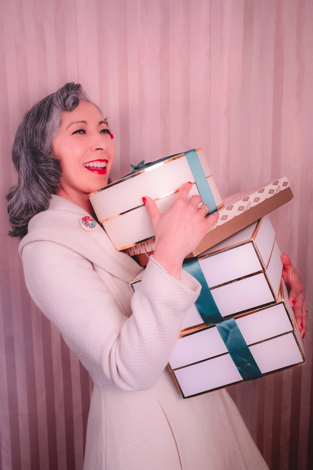 A woman with gray hair and red lipstick smiles while holding several gift boxes wrapped in white and teal ribbon.
