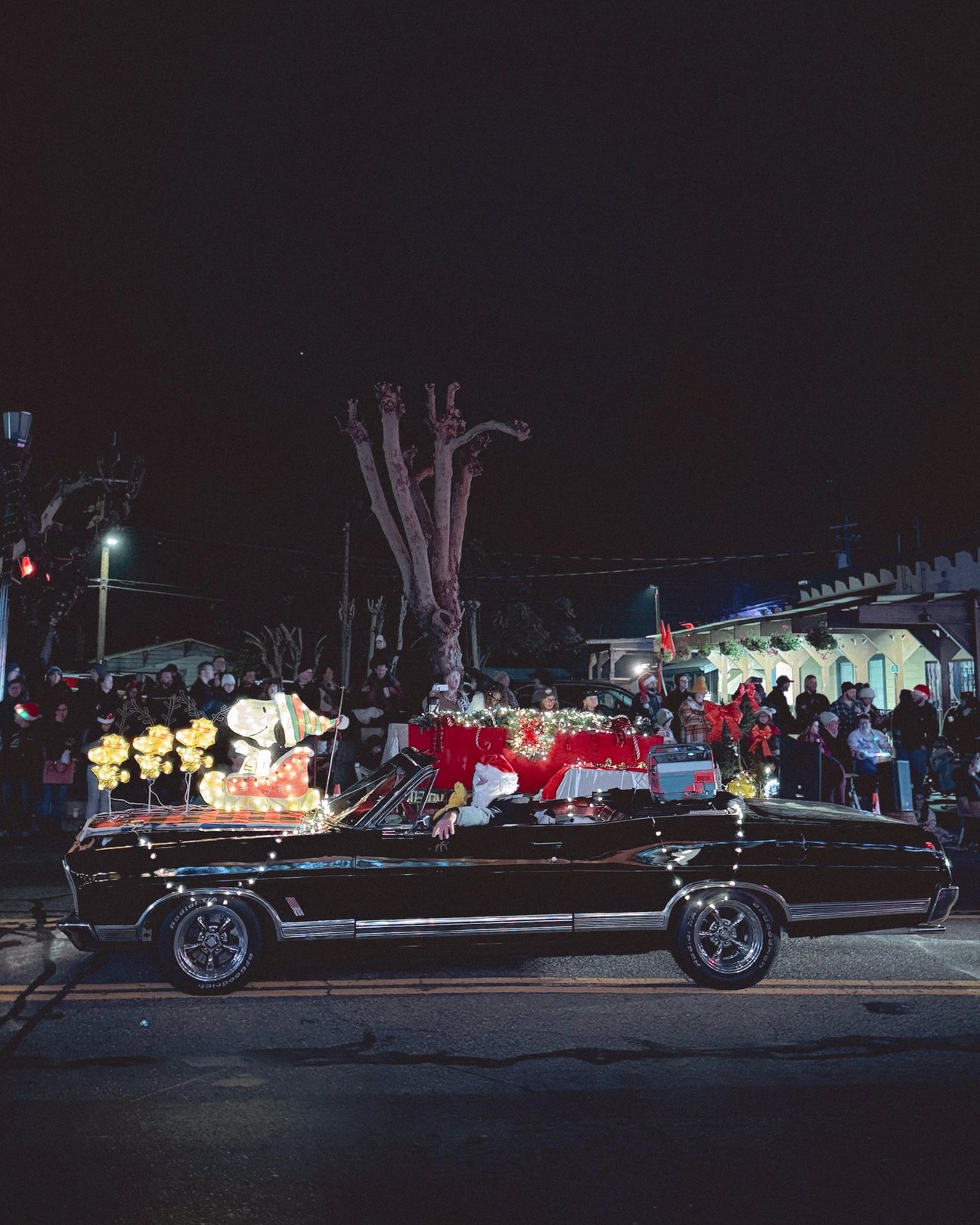 A decorated black convertible car drives past a crowd during a nighttime parade, featuring a festive display with lights and holiday decorations.
