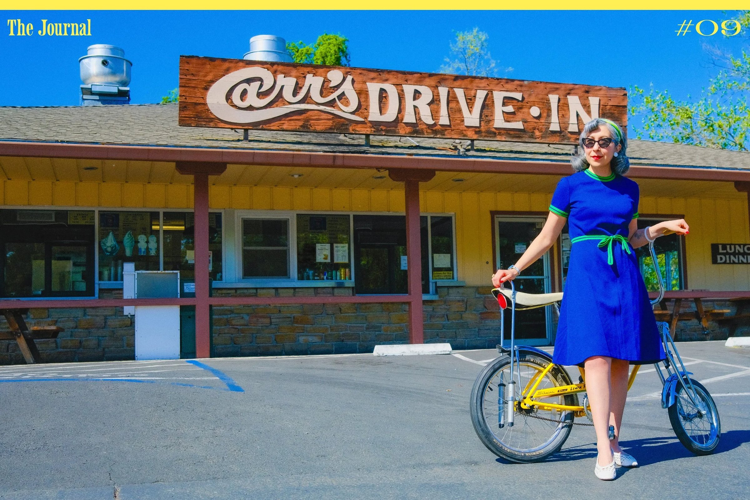 A woman in a blue dress stands beside a yellow bicycle in front of Carr's Drive-In restaurant.