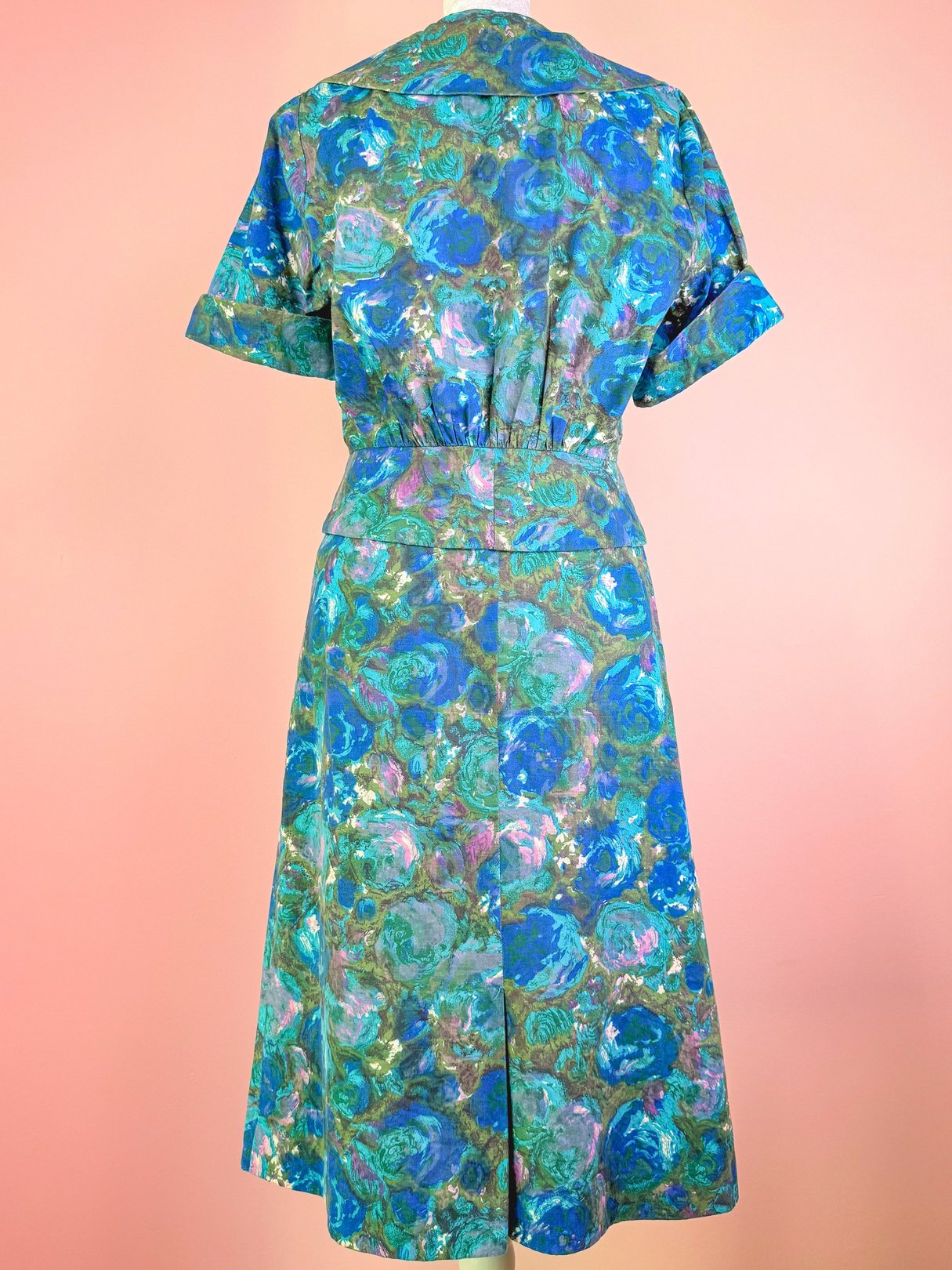 A blue 1950s women's skirt set featuring a floral design and a cinched waist, displayed against a pink background.