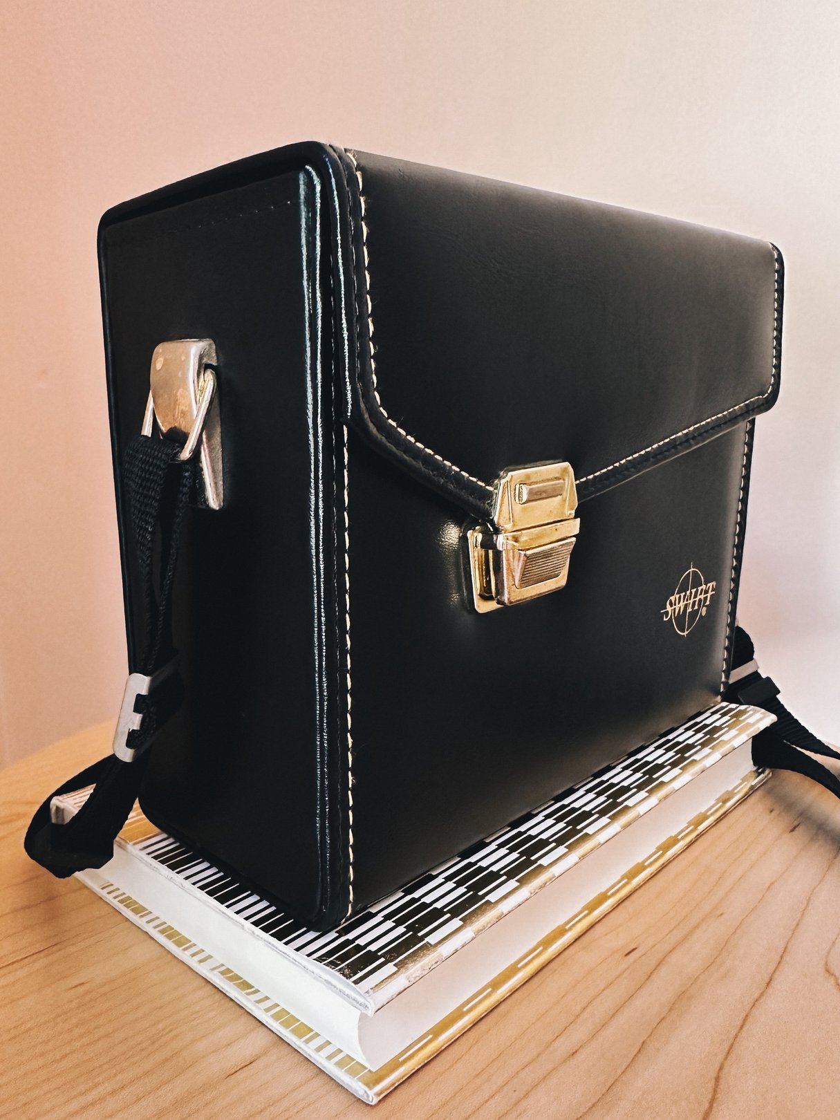 The Swift Adventure Club Camera Bag is a 1970s-style black bag with a gold clasp, featuring a shoulder strap and elegant stitching, displayed on a stack of books.