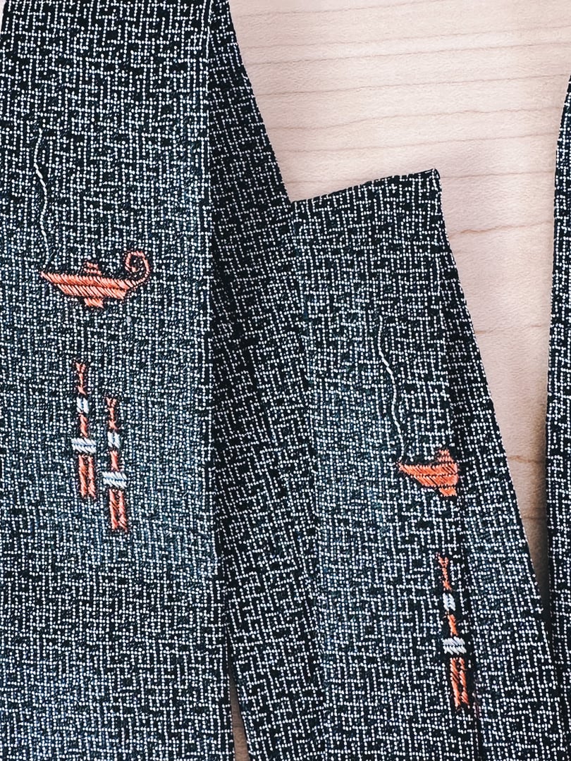 Skinny tie featuring a black textured fabric with embroidered orange genie lamps, inspired by the 1950s style.