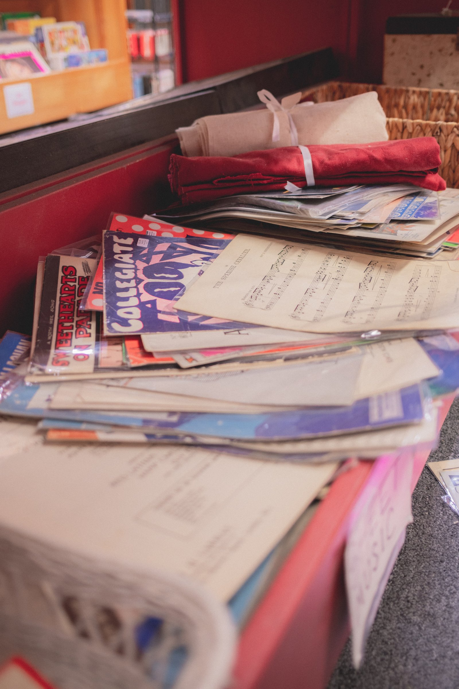 Sheet Music Stories gallery image: A stack of various sheet music and music-related publications is placed on a counter, with a basket and fabric items in the background.