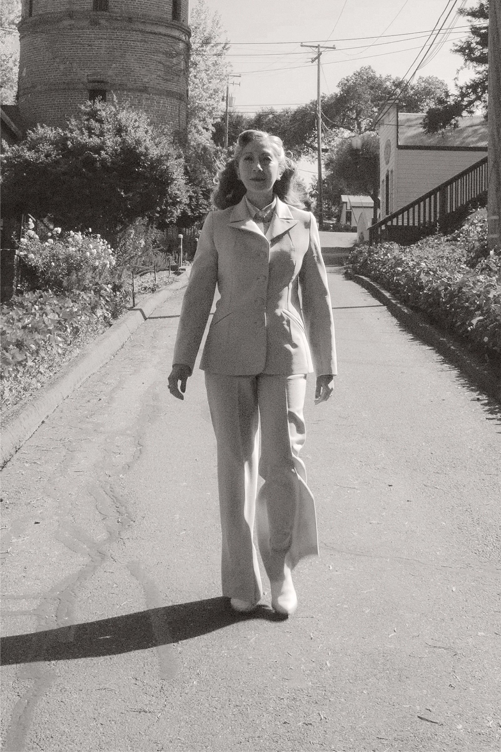 A woman in a light-colored suit walks confidently down a tree-lined street.