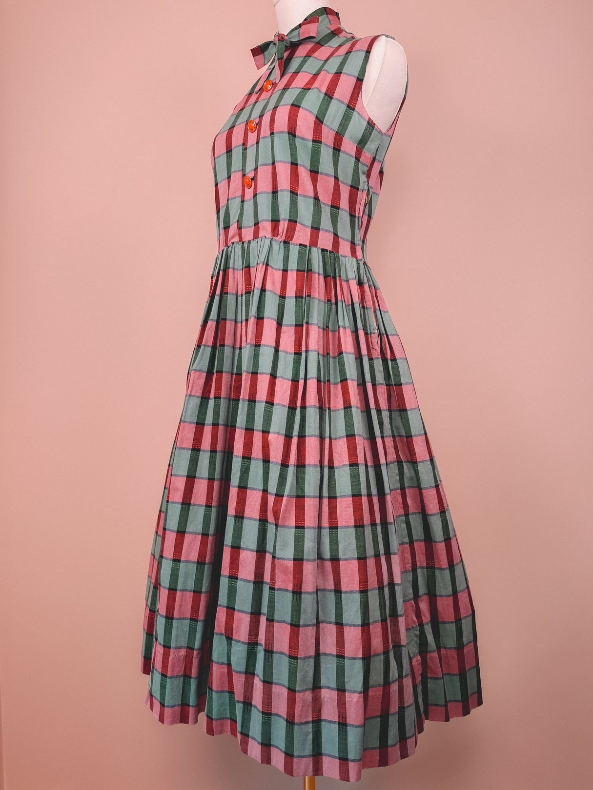 A 1950s women’s plaid dress featuring a sleeveless design with a collar and red buttons, showcasing a pattern of red, green, and pink squares.