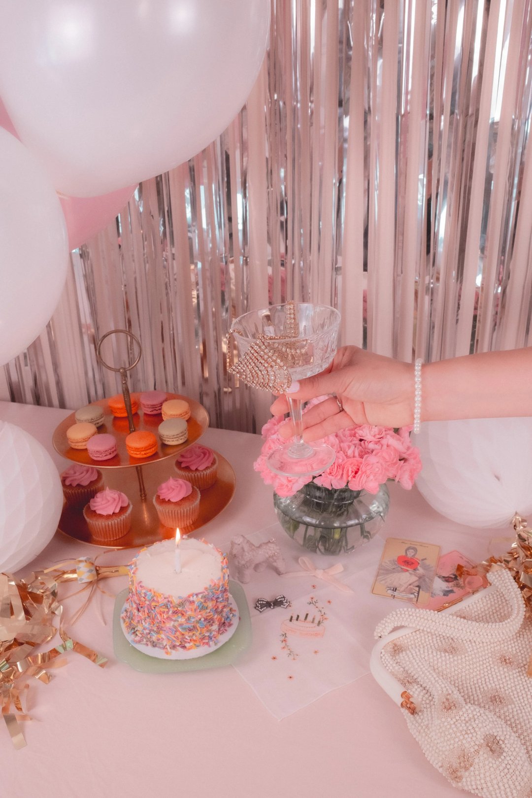 A hand holds a decorated glass beside a vase of pink carnations, a cake with sprinkles, and an array of colorful cupcakes on a table adorned with party decorations.