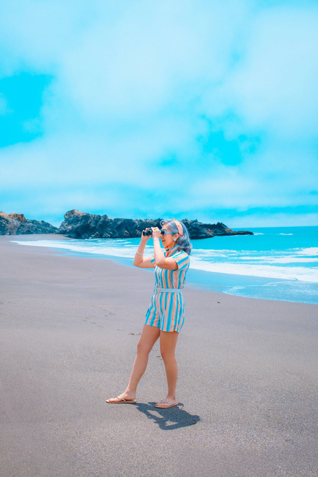 A person in a striped outfit stands on a beach, holding a camera and taking photos of the ocean.