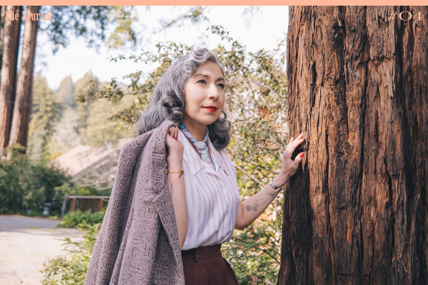 A woman with gray hair poses beside a tree, wearing a striped blouse and holding a coat over her shoulder in a natural outdoor setting.