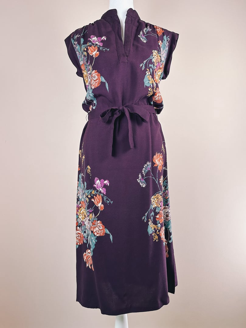 1970s women's purple dress featuring floral patterns and a tied waist.