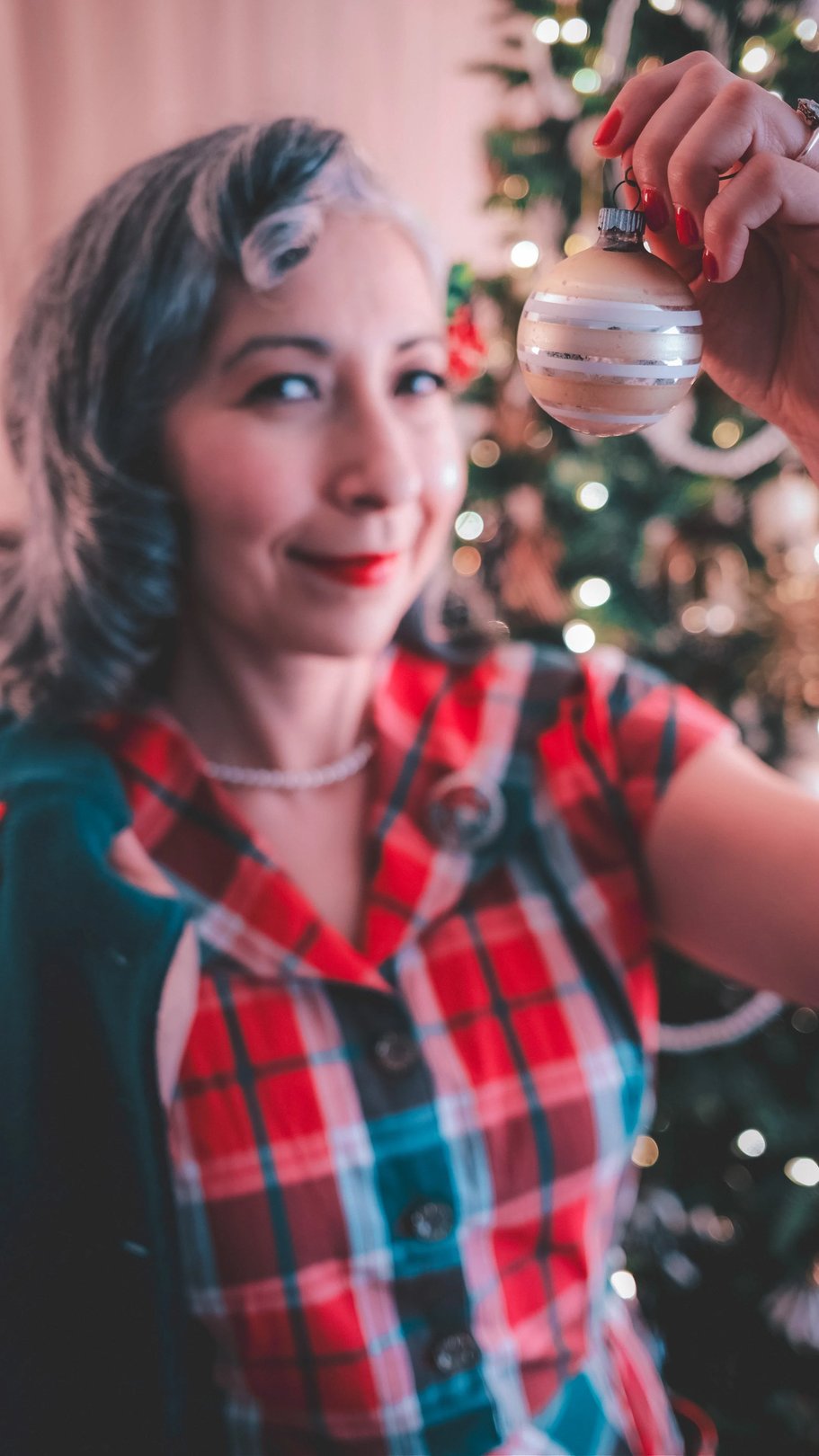 A woman in a plaid dress smiles while holding a vintage Christmas ornament in front of a decorated tree.
