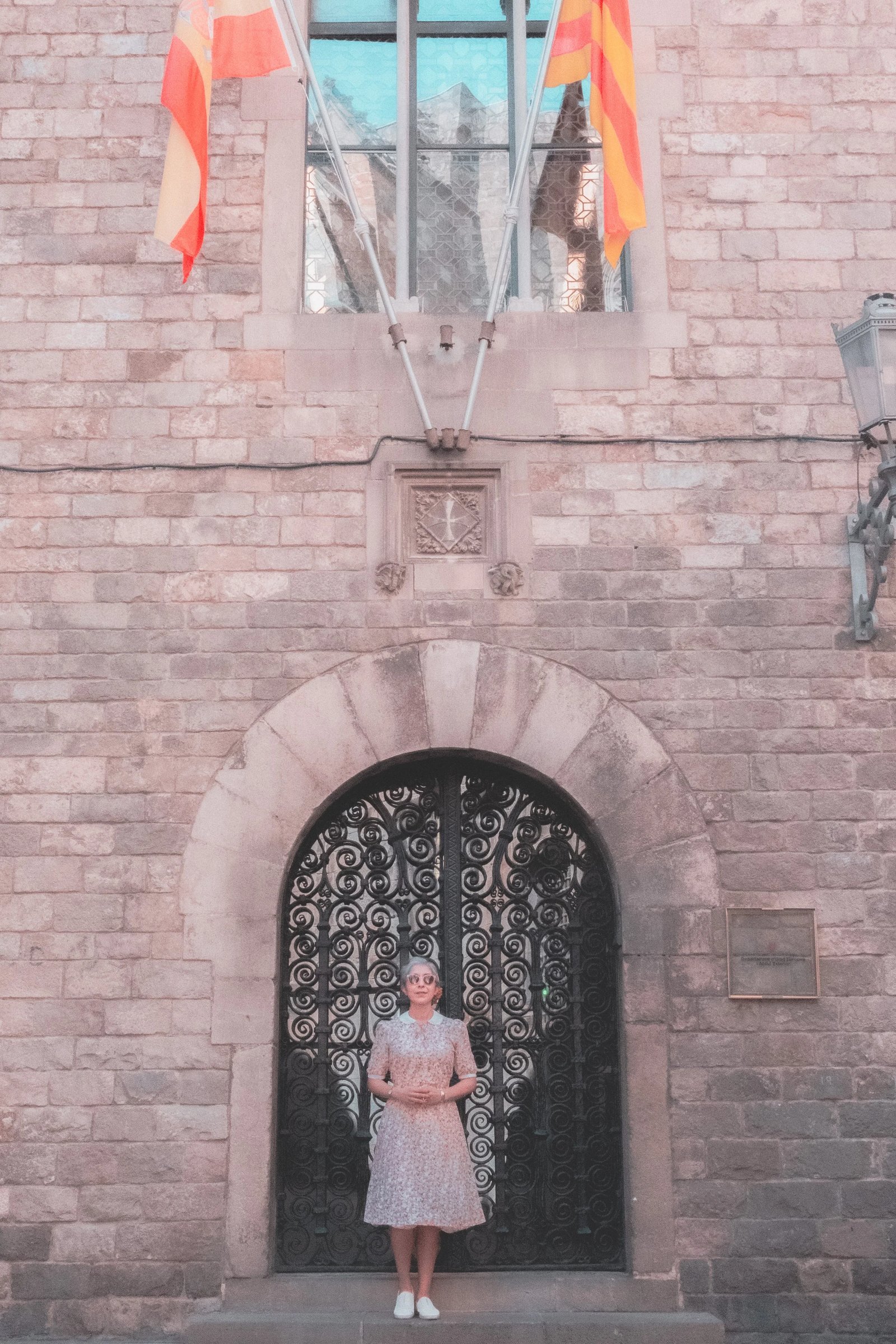 A woman in a floral dress stands in front of an ornate stone archway with decorative iron gates and flags above.