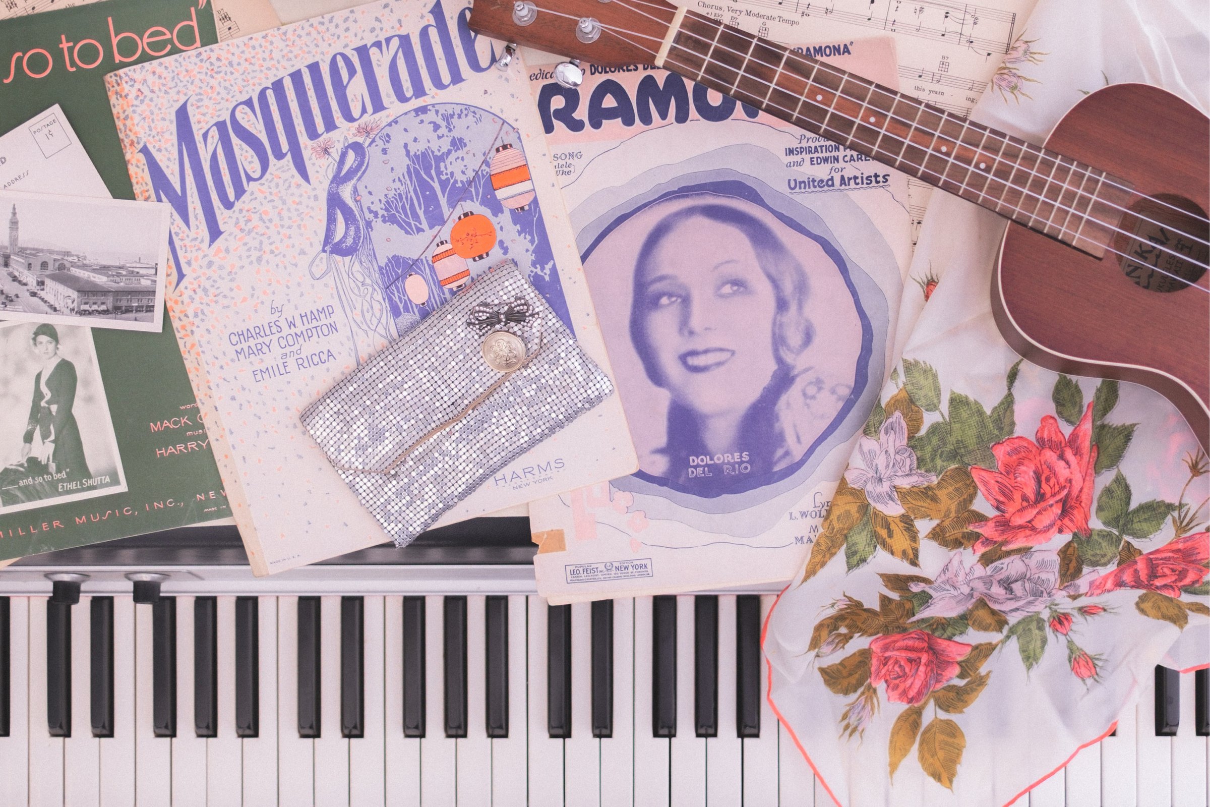 Sheet Music Stories gallery image: A flat lay featuring vintage sheet music, a ukulele, a floral scarf, and a small silver clutch arranged on a piano keyboard.