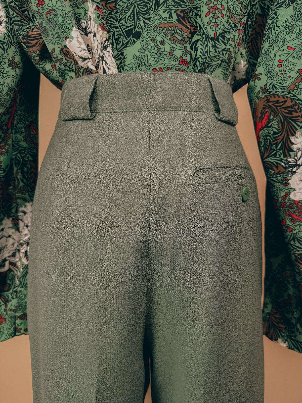 Sage green pleated trousers with a high waist and a back pocket, styled with a floral blouse.