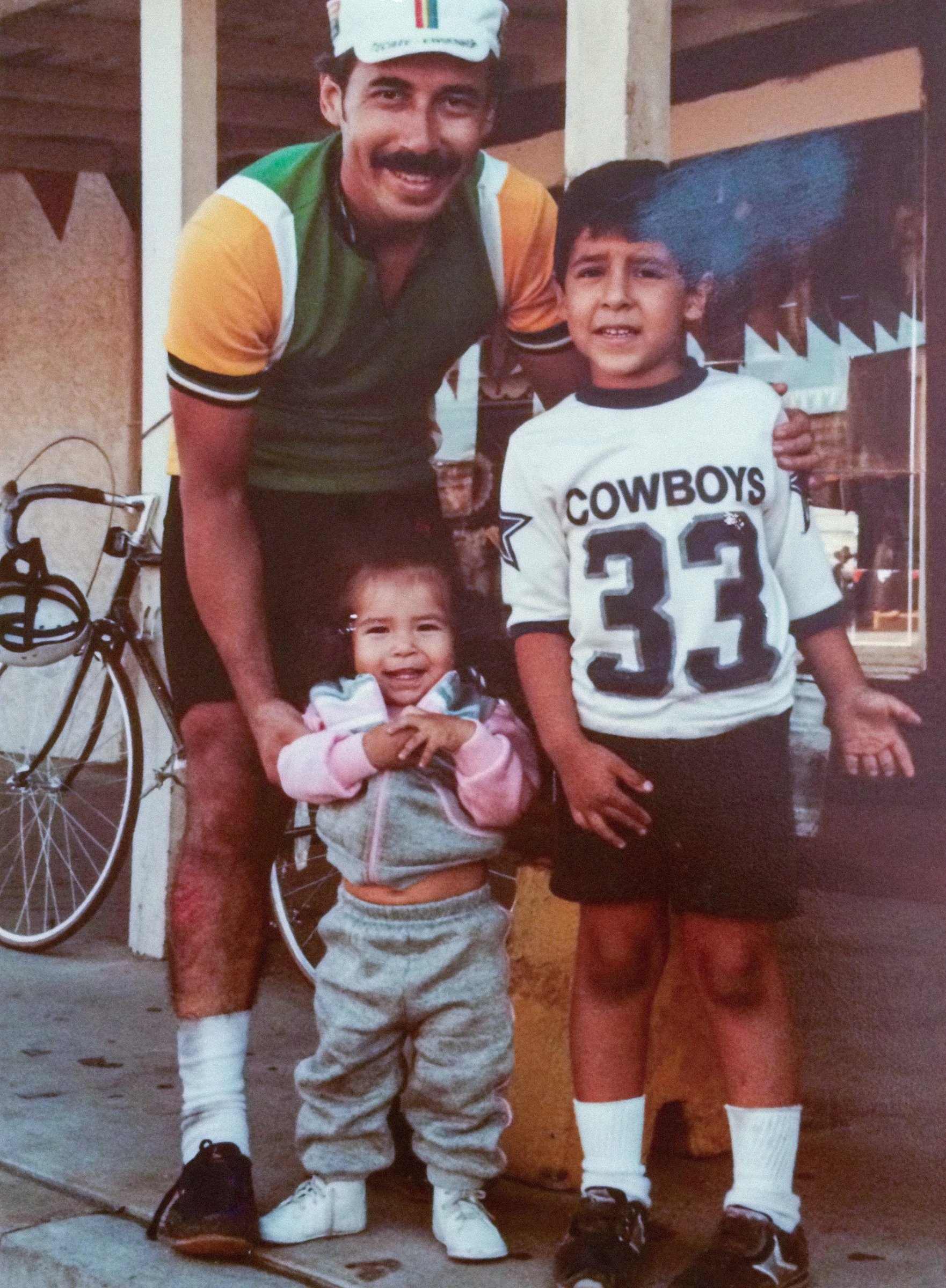 A man with a mustache wearing a cycling helmet and colorful jersey stands beside two children, one toddler in a gray tracksuit and one boy in a Cowboys shirt, at a bicycle shop.