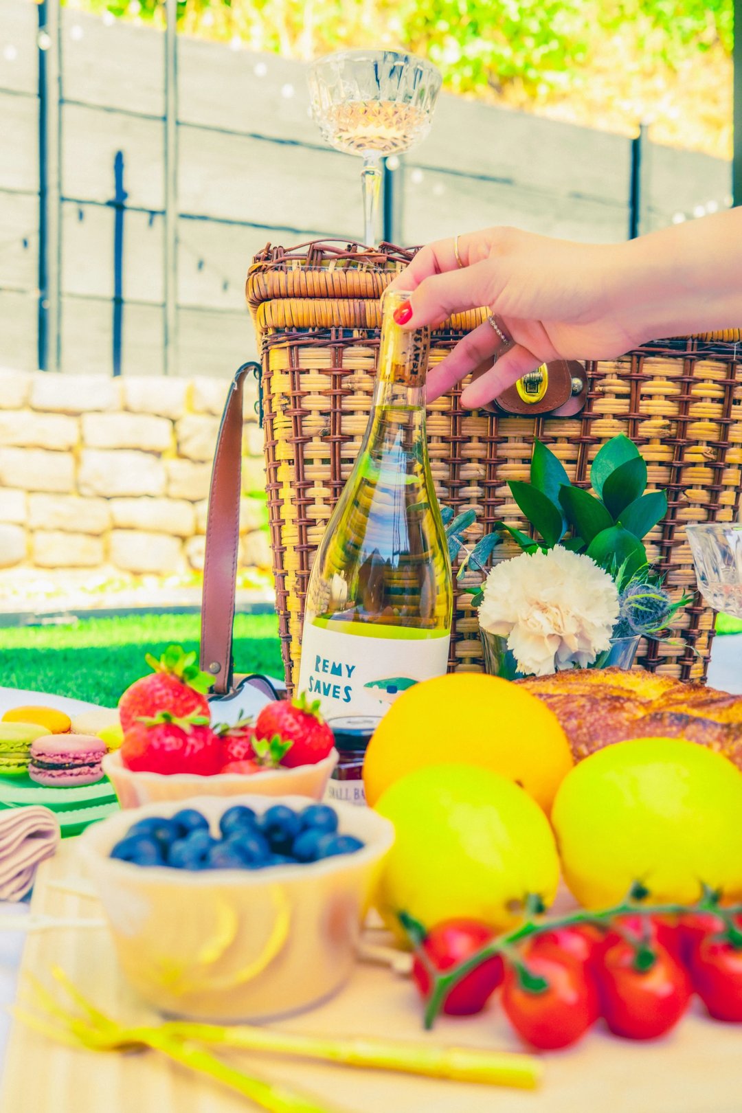 A hand reaches for a bottle of white wine beside a picnic basket, surrounded by fresh fruits, pastries, and a flower.