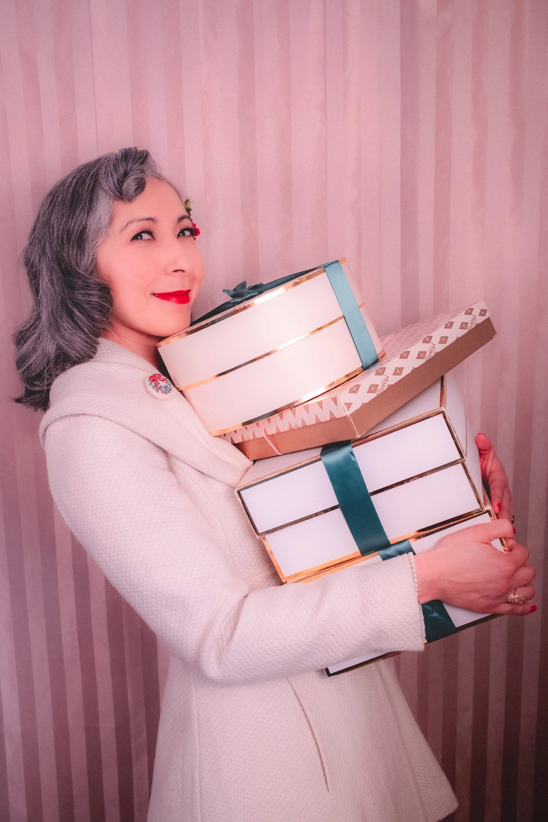 A woman in a white coat holds multiple beautifully wrapped gift boxes with ribbon against a striped pink background.