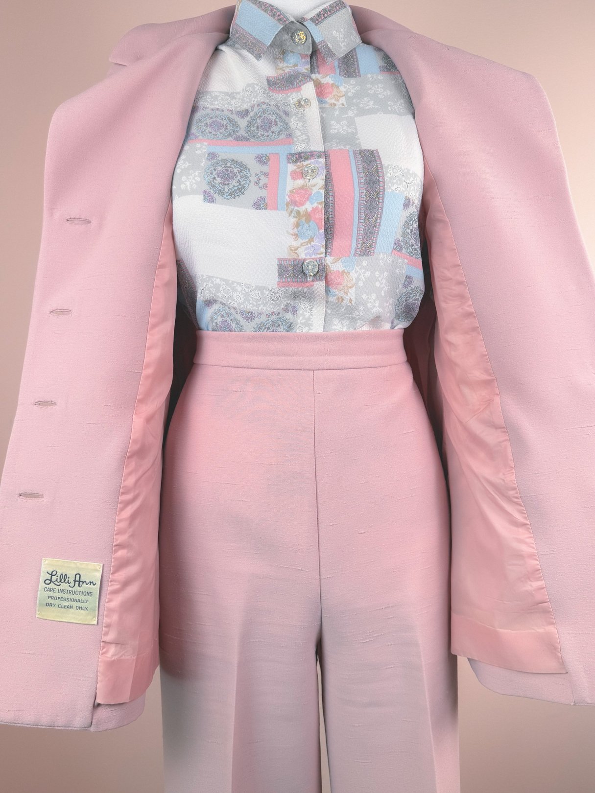 A blush pink three-piece women's suit from Lilli Ann, featuring a matching jacket, blouse with a colorful patterned front, and tailored trousers, typical of 1970s style.