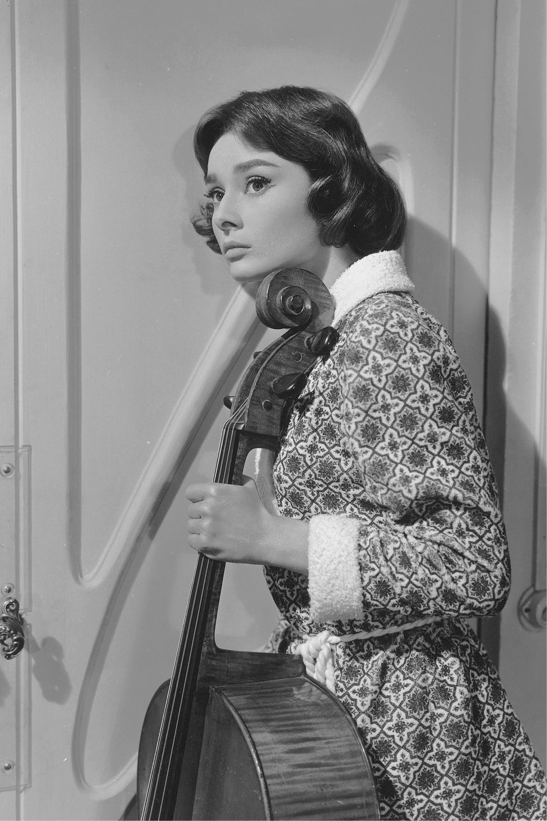 A woman stands with a cello, wearing a patterned robe and smiling softly.