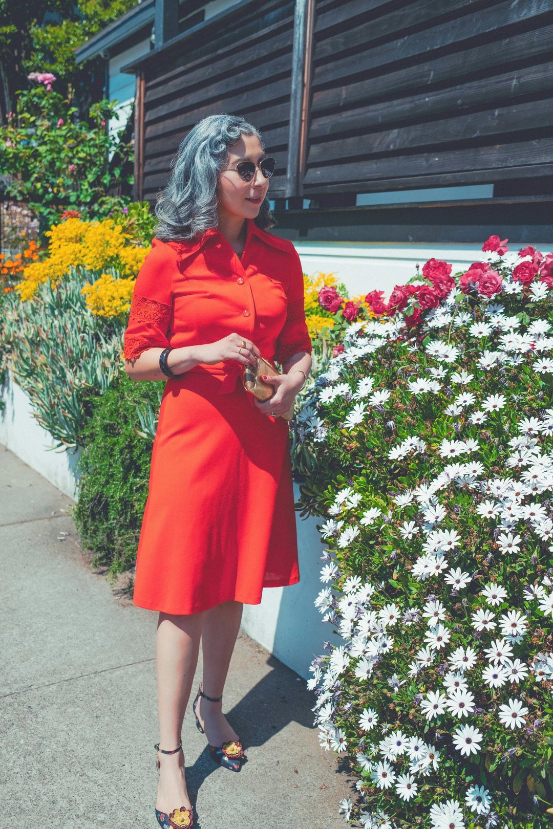 A woman in a red dress with lace detailing stands beside colorful flower beds, wearing sunglasses and holding a small bag.