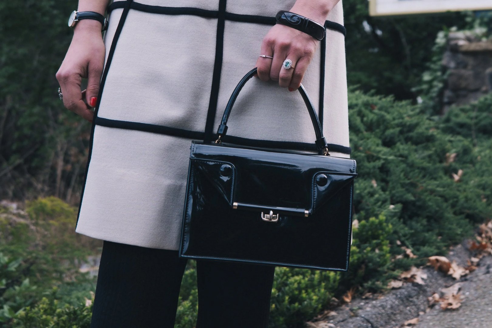 A person holding a glossy black handbag while wearing a light-colored patterned coat.