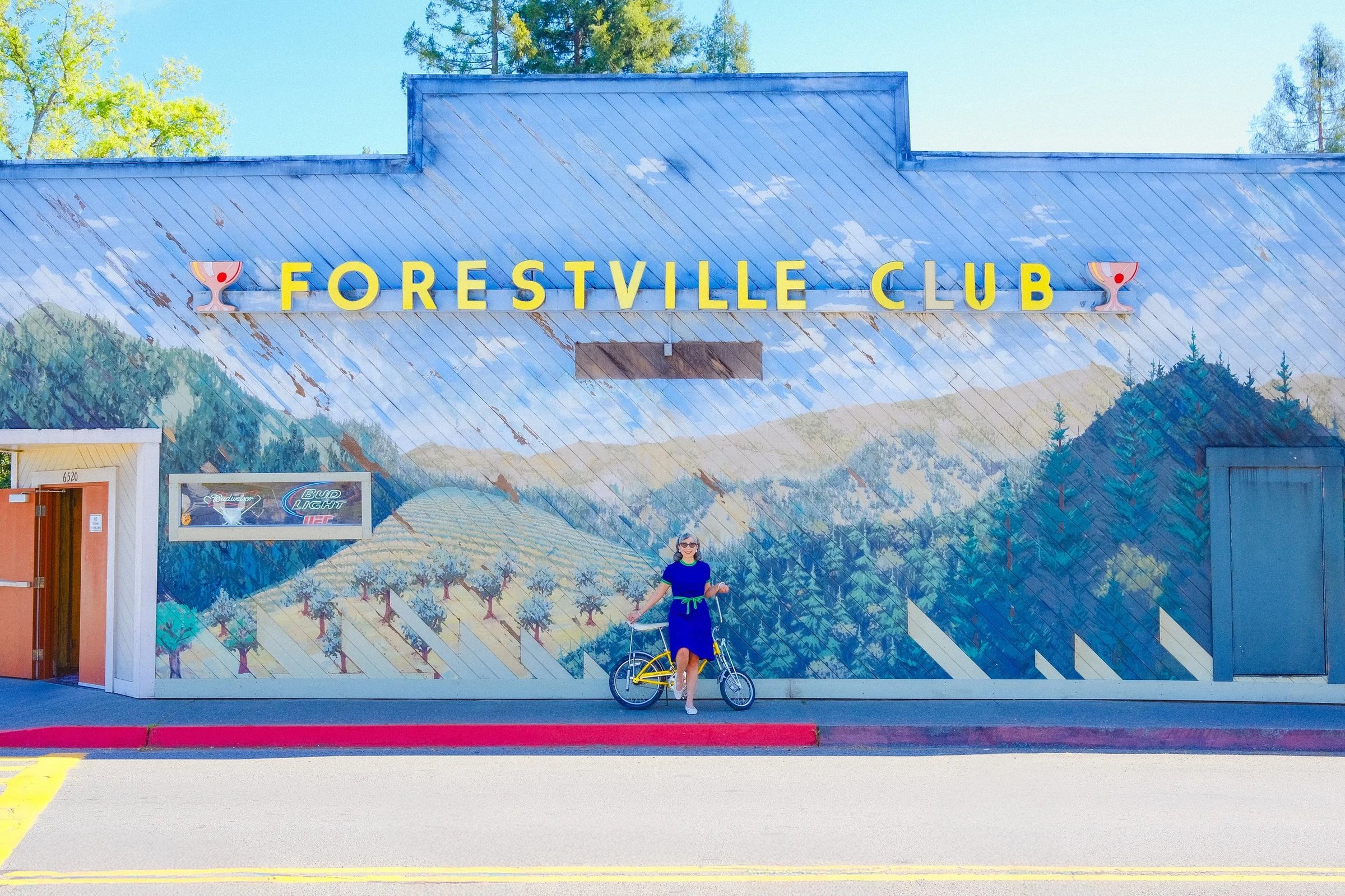 A woman in a blue dress stands beside a yellow bicycle in front of a mural at the Forestville Club.
